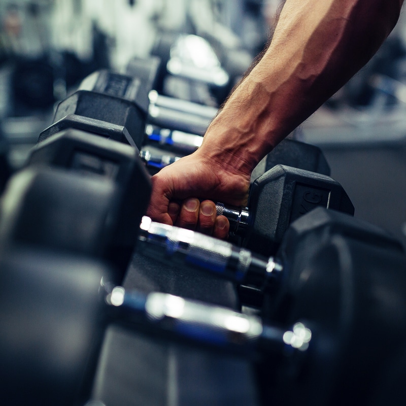 Bras musclé et main saisissant un haltère chromé sur un rack dans une salle de sport. Veines apparentes.