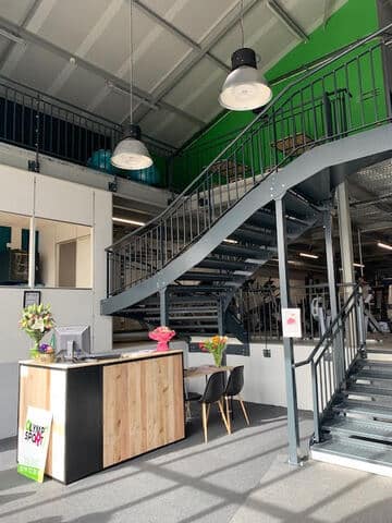 Intérieur moderne d'une salle de sport Olymp Sport avec comptoir d'accueil, grand escalier métallique et équipement de fitness en arrière-plan.