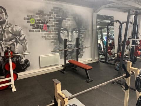 Salle de sport moderne avec mural "OLYMP SPORT" esprit graffiti. Un homme musclé et un visage féminin ornent le mur. Équipement complet: banc de musculation, barres, haltères et miroirs.