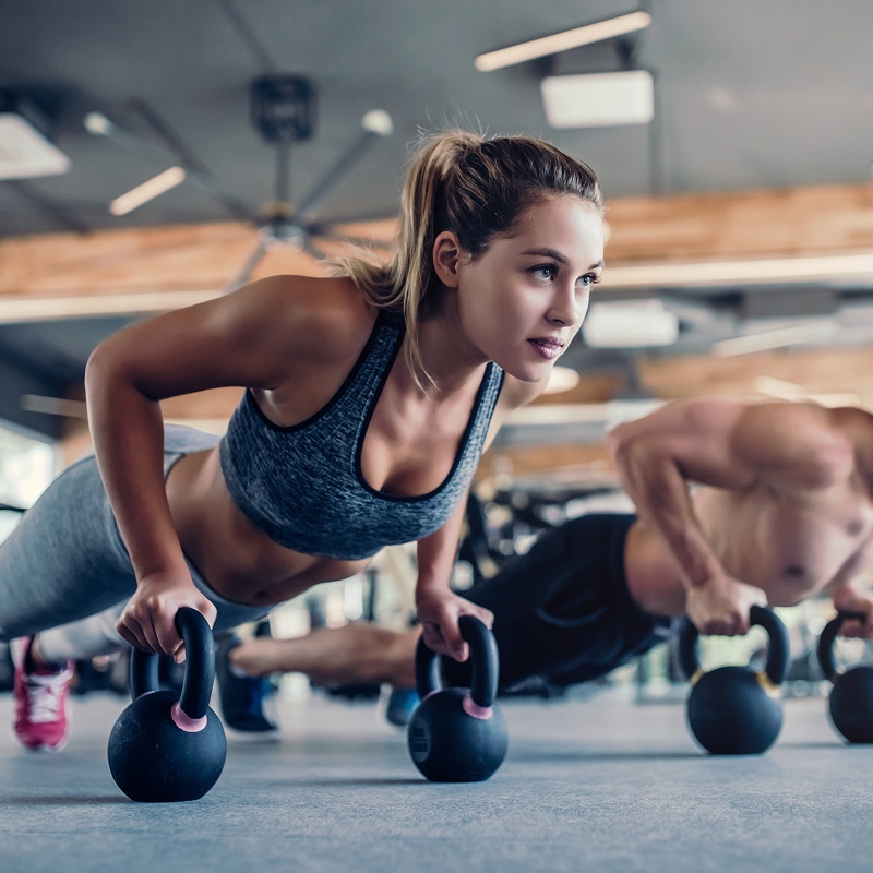 Femme et homme faisant des pompes sur kettlebells en salle de sport. La femme, au premier plan, est concentrée et musclée.