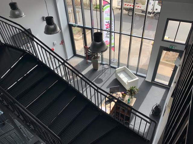 Vue plongeante sur un intérieur de bureau moderne avec escalier métallique noir, suspensions industrielles, canapé blanc et panneau EXIT/SORTIE.