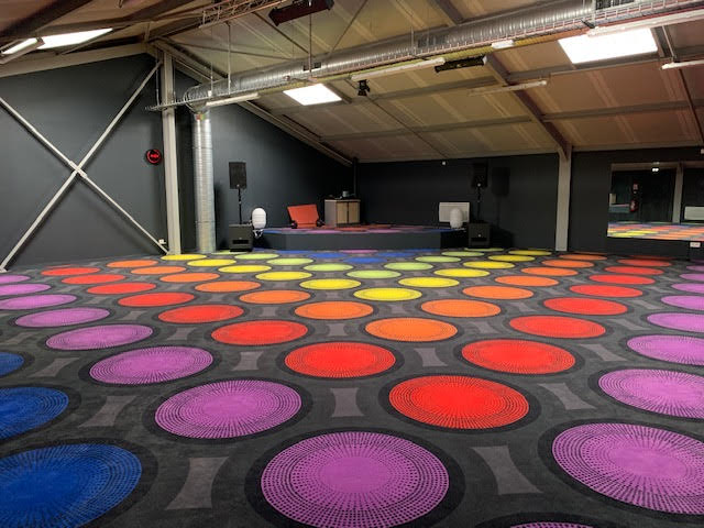 Salle événementielle moderne avec moquette à motifs de cercles multicolores vibrants. Scène équipée de son et lumière à l'arrière.