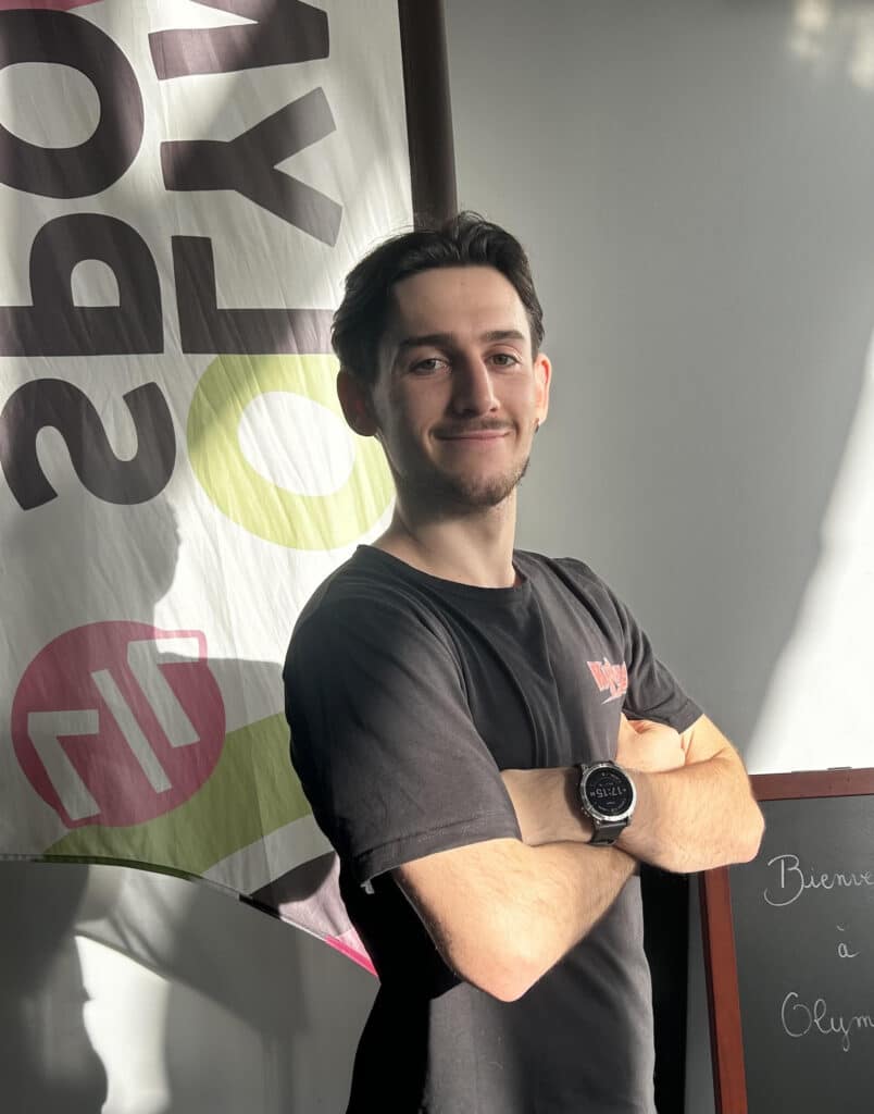 Coach souriant avec montre sport devant bienvenue Olym Jeune homme souriant avec les bras croisés, portant une montre connectée, devant un panneau à texte renversé et un tableau noir "Bienvenue à Olym".