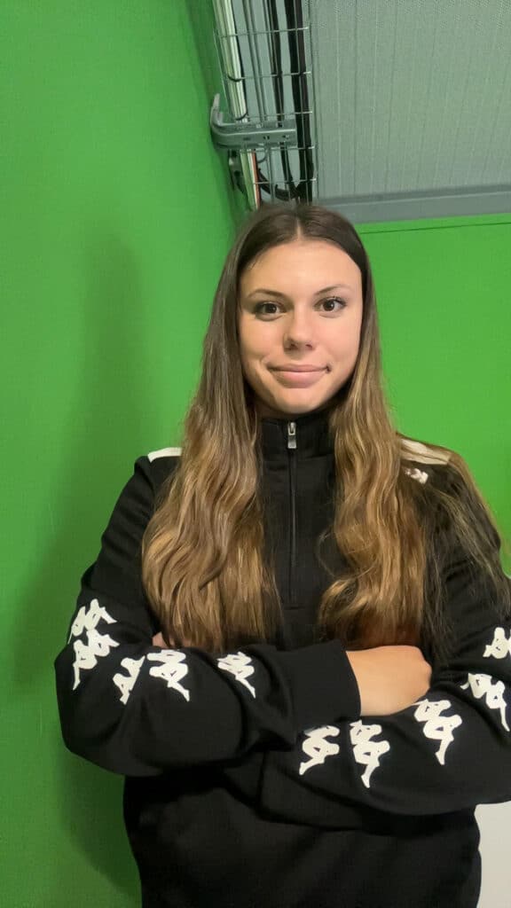 Sportive Kappa avec survêtement noir fond vert Jeune femme souriante vêtue d'un survêtement noir Kappa avec logo sur les manches devant un fond vert.